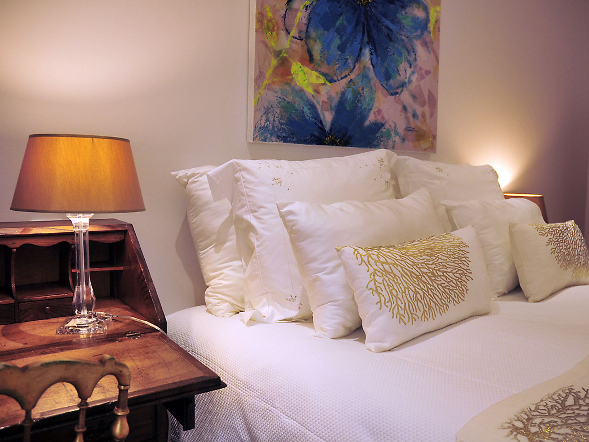 Una cama bien hecha con ropa de cama blanca y detalles en pan de oro, una mesita de noche de madera con una lámpara y un cuadro sobre la cama.