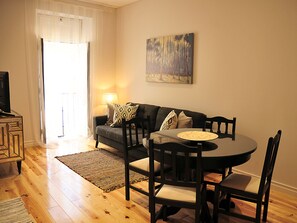 Ein gemütliches Wohnzimmer mit einem dunkelgrauen Sofa, einem runden Esstisch und einem Fernseher auf einer Holzkommode.