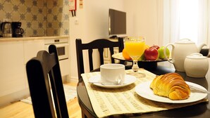 A breakfast table with croissant, fruit, and juice, a coffee maker, and a TV in the background. - Fully equipped air-conditioned apartment in central Setúbal (Setúbal)