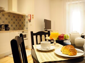 A breakfast table with croissant, fruit, and juice, a coffee maker, and a TV in the background. - Fully equipped air-conditioned apartment in central Setúbal (Setúbal)