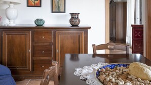 A dining area with a wooden table, chairs, and a plate of nuts. There are paintings on the wall and a lamp on the sideboard. - Apartment in two-room villa called Lipari 4/5 beds (Reitano)