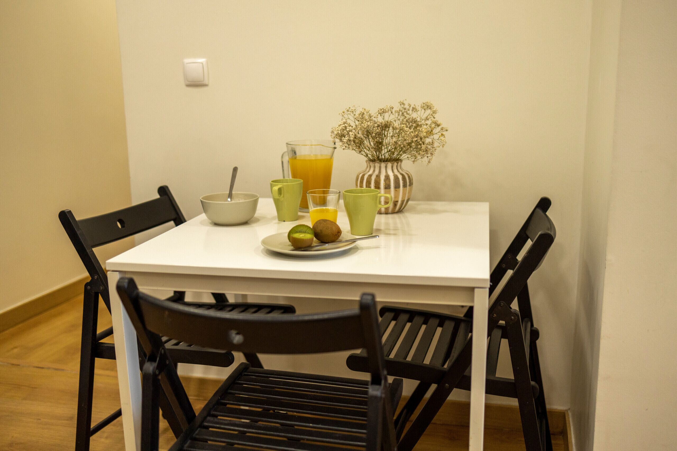Ett matbord med stolar, en kanna med apelsinjuice, en skål, en tallrik med två avokadon och en vas med blommor.
