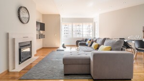 A modern living room with a fireplace, a grey sofa, and a large window providing natural light. - Cascais Park (Cascais)