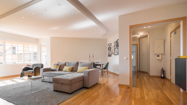 A spacious living area with a sectional sofa, a coffee table, and a dining table. There is a wooden floor and a large window providing natural light. - Cascais Park (Cascais)
