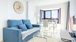 A modern living room with a blue sofa, a glass coffee table, a dining area with a white table and chairs, and a large window with curtains. - Torre Mar 20-D (Benidorm)