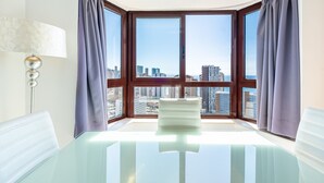 A modern room with a glass table, a white chair, and a view of a cityscape through large windows with curtains. - Torre Mar 20-D (Benidorm)
