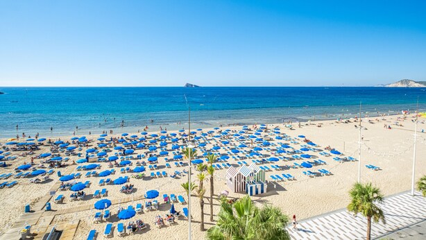 A beach with numerous blue umbrellas and sun loungers, a clear blue sky, and a calm sea. - Torre Mar 20-D (Benidorm)