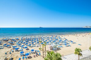 Una platja amb nombrosos paraigües blaus i gandules, un cel blau clar i un mar tranquil.