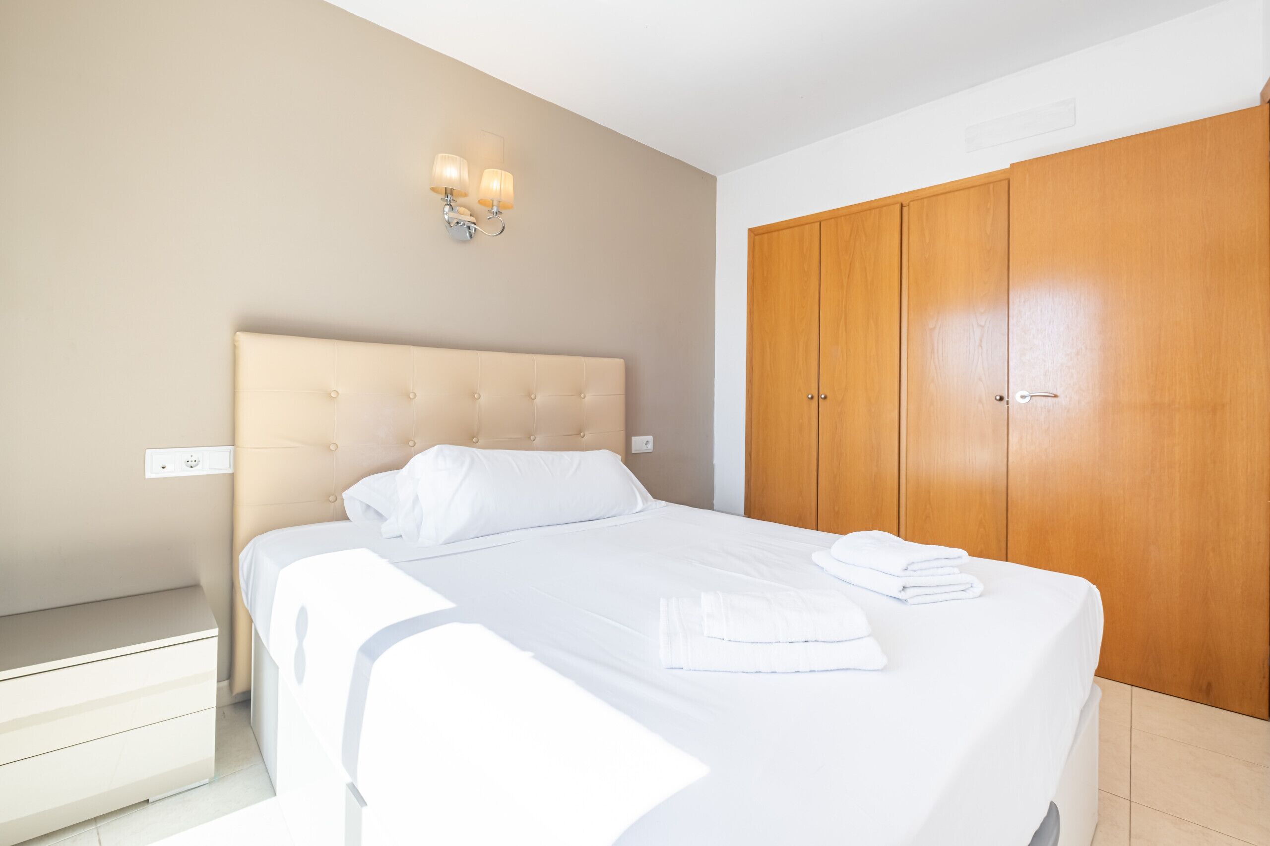 A neatly made bed with white linens and a headboard, a wooden wardrobe, and a wall-mounted light fixture.