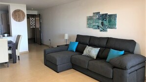 Een moderne woonkamer met een modulaire bank, een eethoek met een tafel en stoelen, en een keuken op de achtergrond.