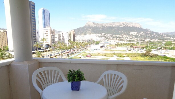 Un balcon avec une table ronde et quatre chaises, d’où l’on peut admirer un paysage urbain avec des bâtiments et une montagne à l’horizon.