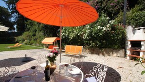An outdoor dining area with a table set for four, a wine bottle, and a large orange umbrella. - Cascais Downtown, Villa With Pool and Garden (Cascais)