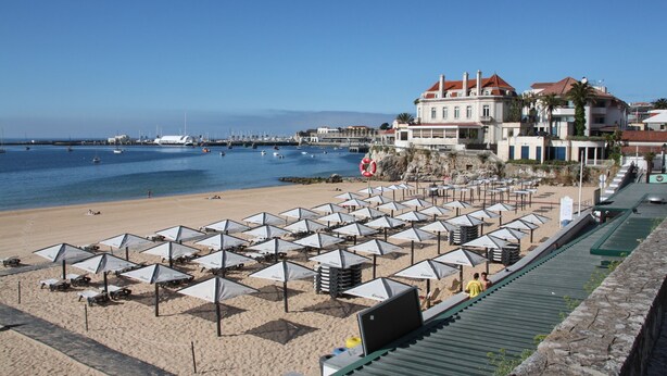 A beach with numerous umbrellas and lounge chairs, a large building with a red roof, and a clear blue sky. - Cascais Downtown, Villa With Pool and Garden (Cascais)
