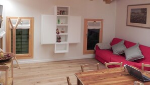 Ein gemütliches Zimmer mit Holzboden, einem roten Sofa mit Kissen, einem Holztisch mit einem Laptop und einer weißen Regaleinrichtung an der Wand.