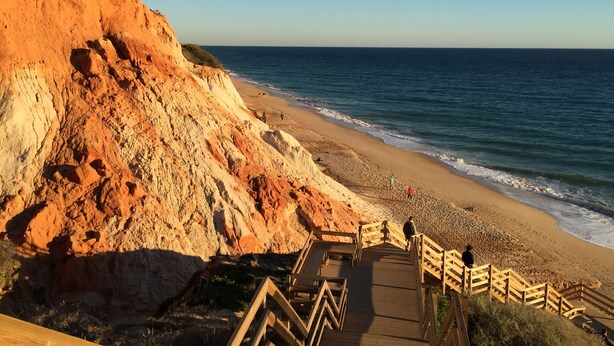 Un paysage côtier avec un escalier en bois descendant vers une plage de sable, offrant une vue sur une falaise aux formations rocheuses de couleur rouge-brun.