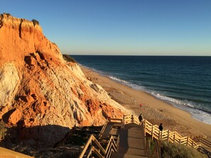 Eine Küstenlandschaft mit einer Holztreppe, die zu einem Sandstrand führt, mit Blick auf eine Klippe und rötlich-braune Felsformationen.