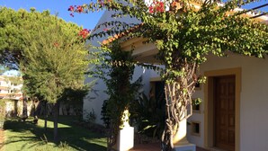 Une maison dotée d’un patio couvert, d’une porte en bois et d’un jardin où poussent diverses plantes et arbres.