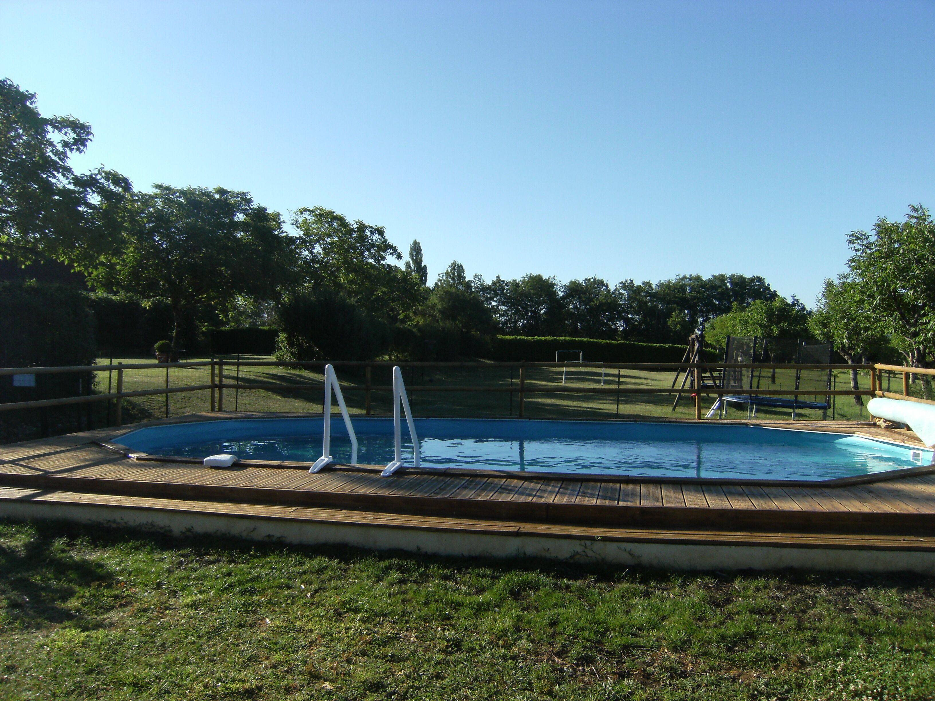 Uma piscina retangular com deck de madeira, rodeada por uma área cercada e árvores ao fundo.