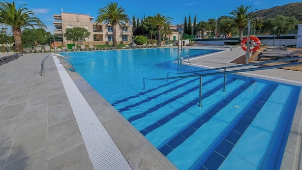 En swimmingpool med kristallklart blått vatten, omgiven av palmer och en modern byggnad i bakgrunden.