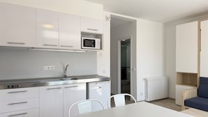 Ett modernt kök med vita skåp, en mikrovågsugn och en diskho. I det intilliggande vardagsrummet finns ett matbord med stolar, ett element och en blå soffa.