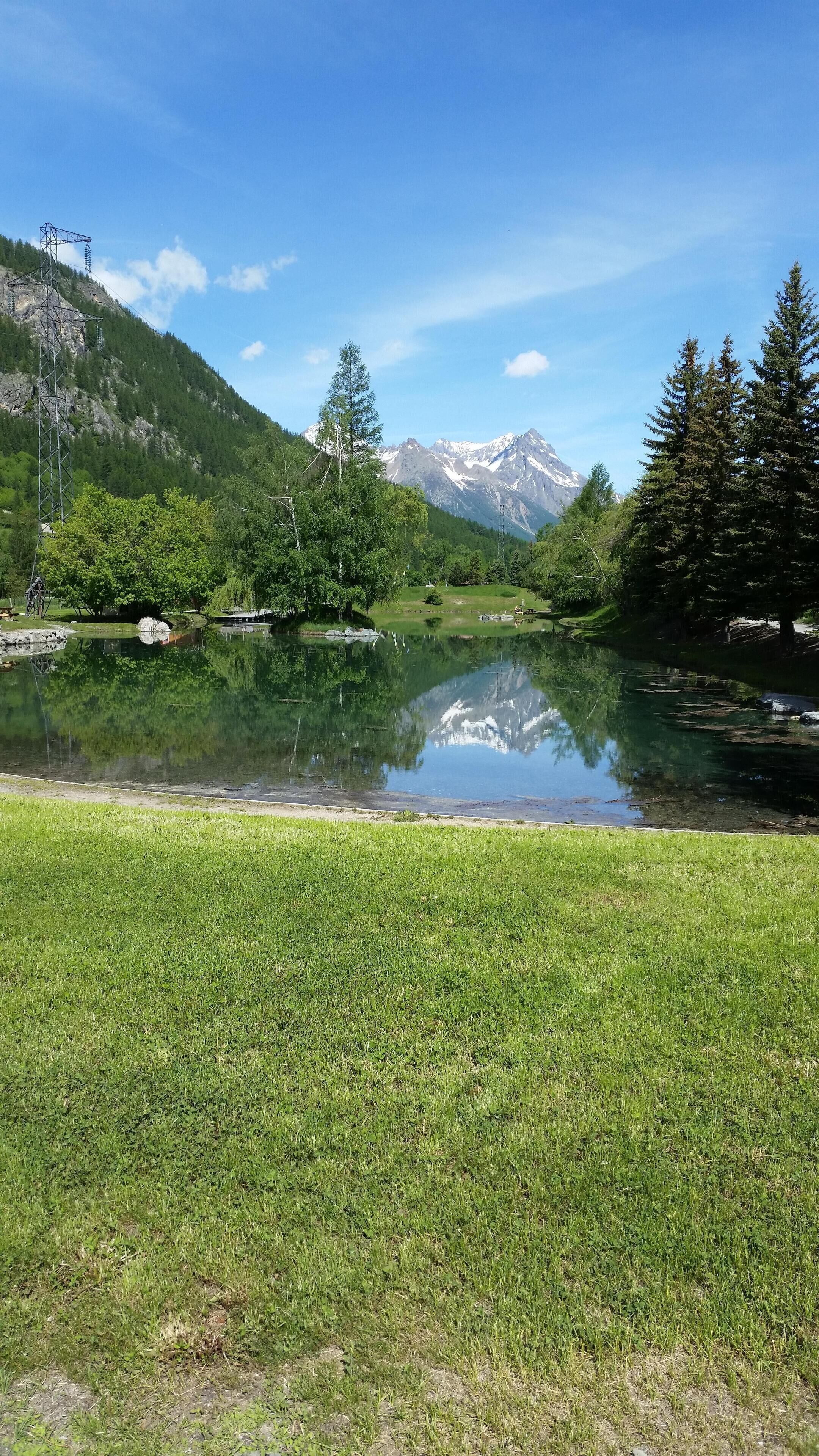 Ein klarer See mit Bergen im Hintergrund, umgeben von grünen Bäumen und Gras.