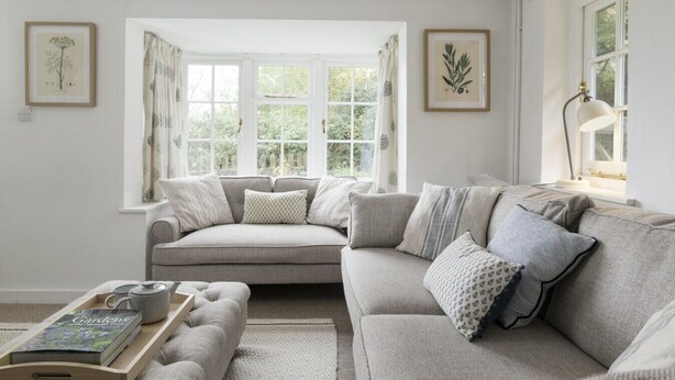 A cozy living room with a grey sofa, a small table with a book and a teapot, and two framed botanical illustrations on the walls.