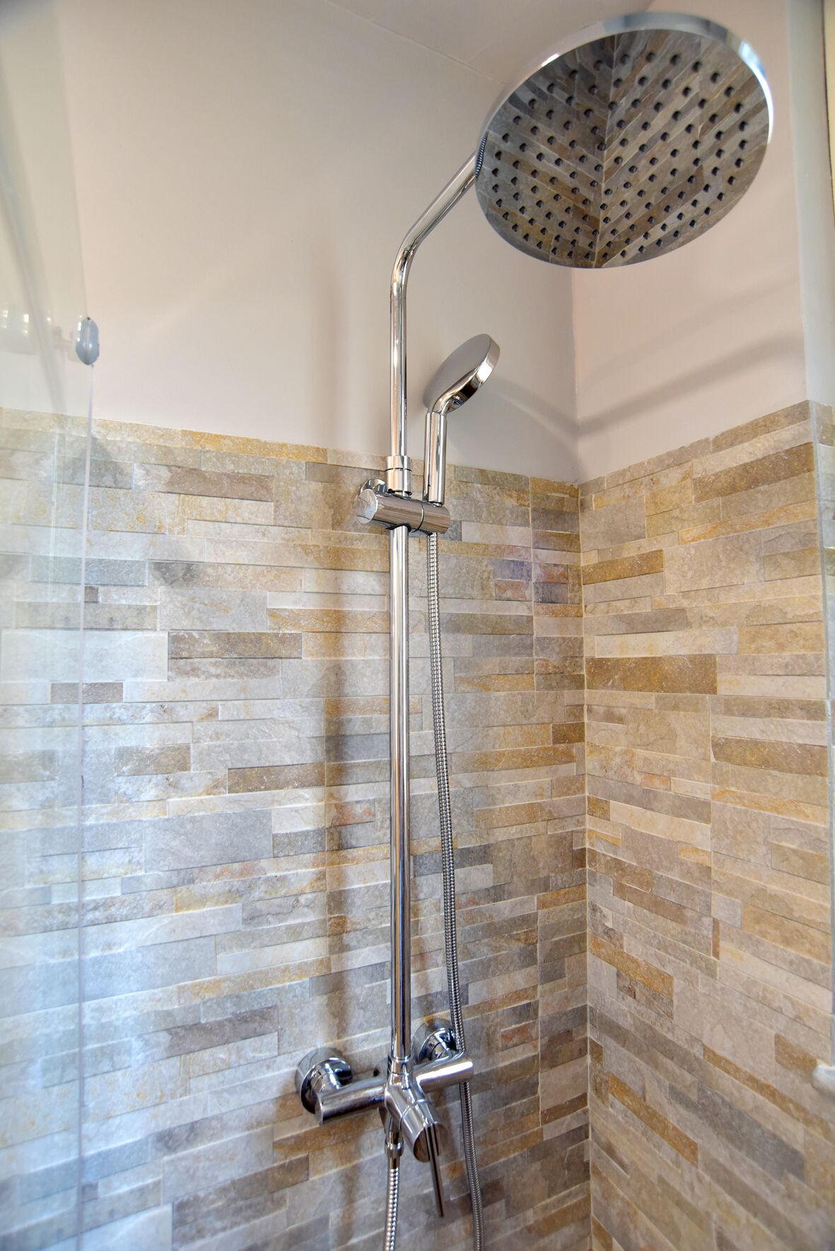 Dusche mit Regendusche und Handbrause, vor einer mit Steinmuster belegten Wand.