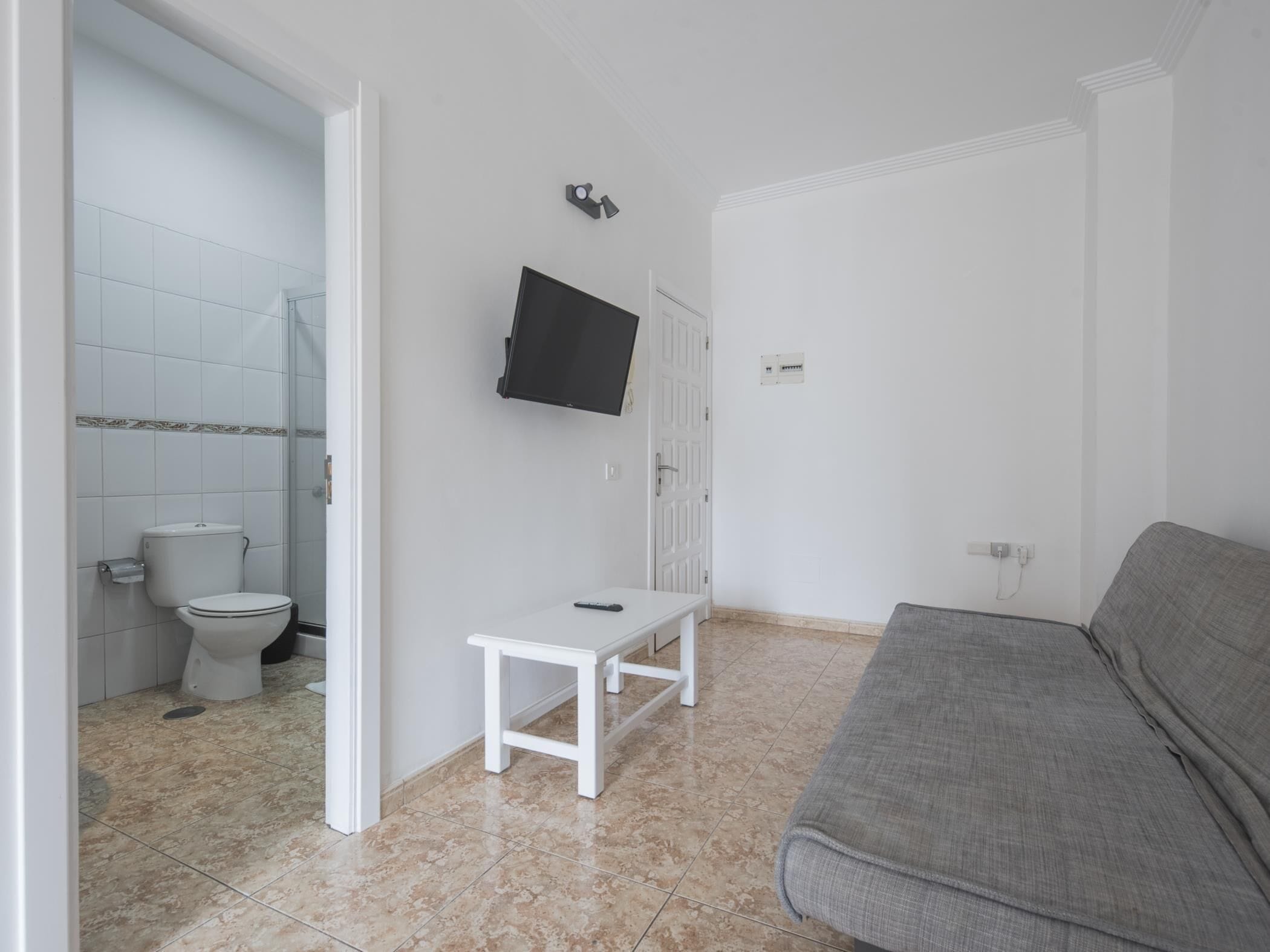 Et lille, rent værelse med toilet, bænk, en grå sofa og et fjernsyn ophængt på væggen.