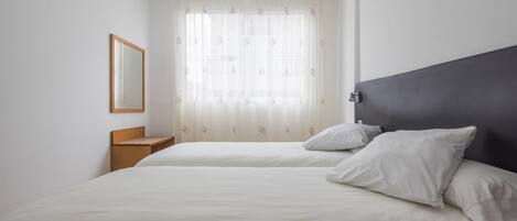 A hotel room with two beds, a wooden nightstand, a mirror, and a window with sheer curtains.