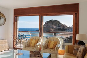 A room with two chairs, a sofa, a glass table, a clock, and a view of the sea and cliffs.
