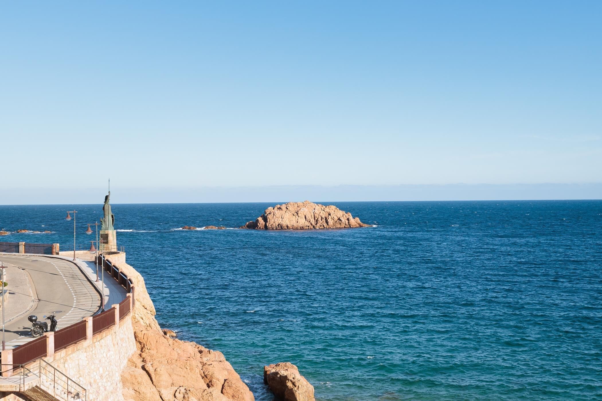 A coastal view with a rocky outcrop, a paved walkway, and a statue.
