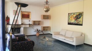 Een woonkamer met een bank, een bijzettafeltje, een vloerlamp en een kroonluchter. Er staan planken met decoratieve objecten en er hangt een schilderij aan de muur.