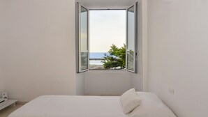 A bedroom with a bed, a white wall, and a window with a view of the sea and a tree.