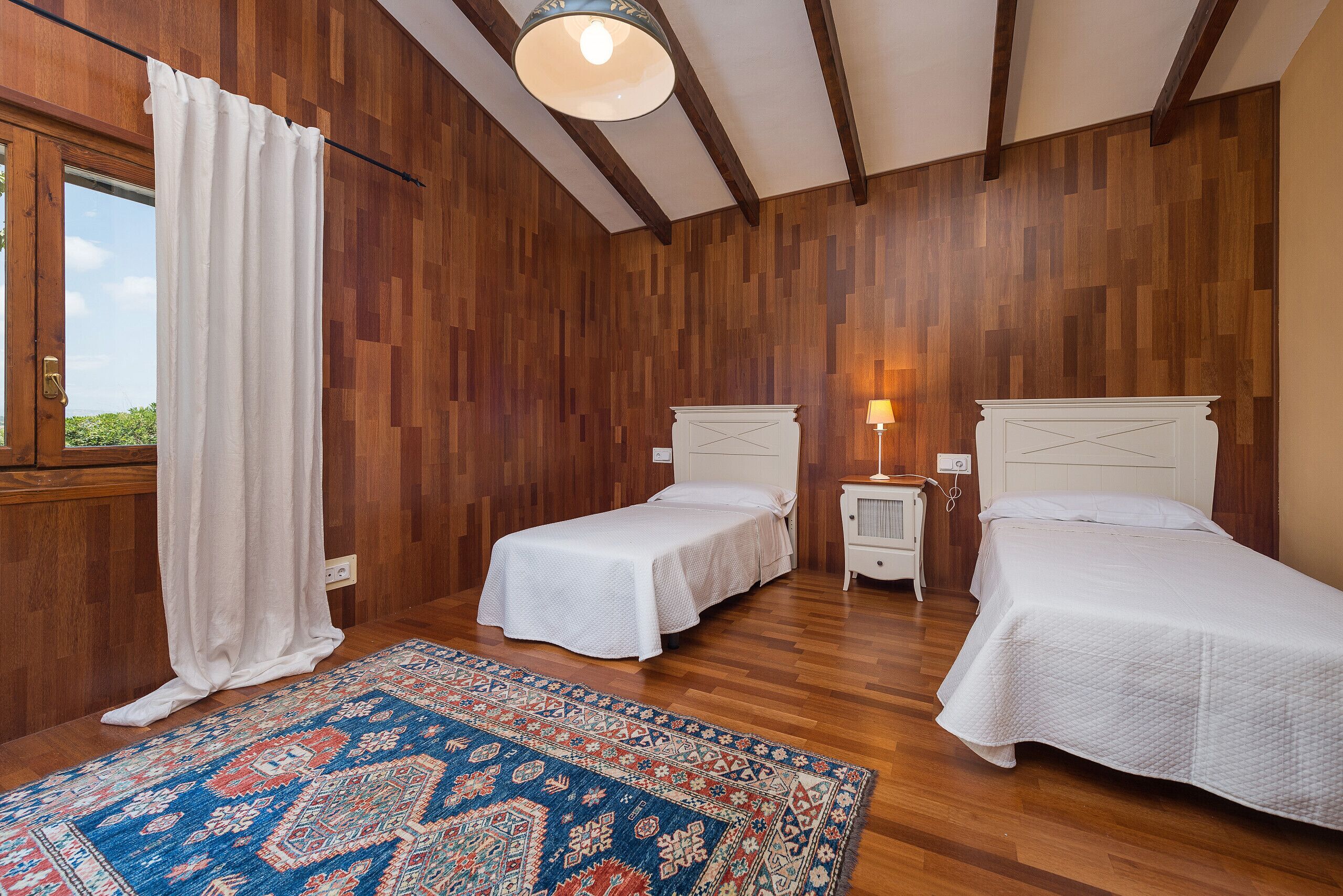 Una habitación con dos camas, una ventana con cortinas, un tapete estampado y una pared de madera.