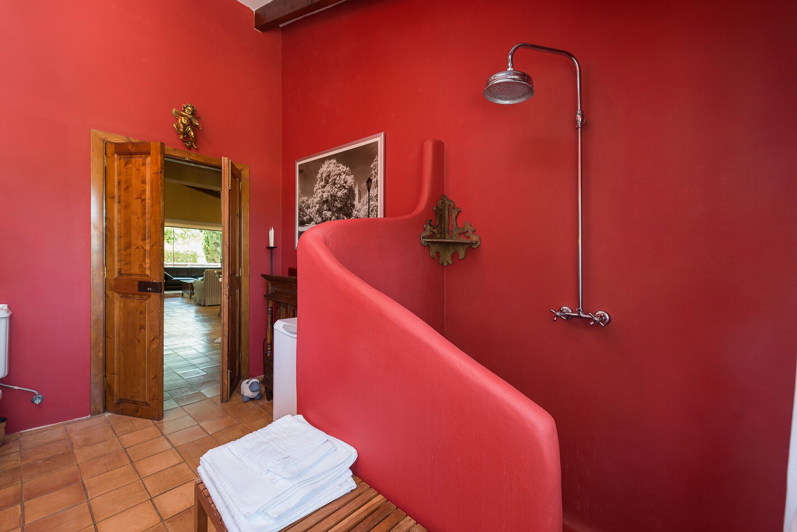 A bathroom with a red wall, a shower, a wooden bench, and a door leading to another room.