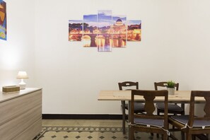 Un coin repas avec une table et des chaises, une œuvre d’art accrochée au mur, une lampe et une petite plante posée sur la table.