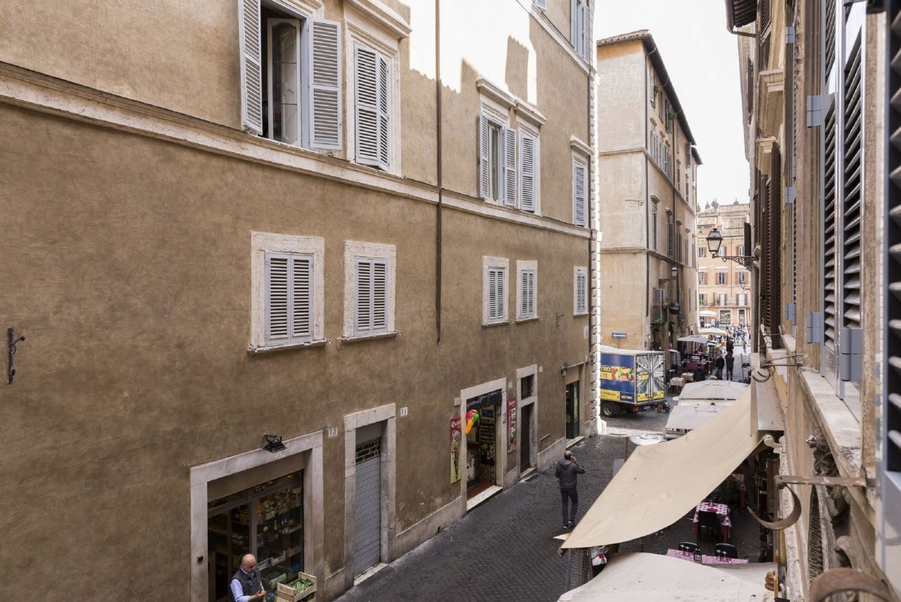 A narrow street with buildings on either side, featuring shutters, a street vendor cart, and a small outdoor seating area.