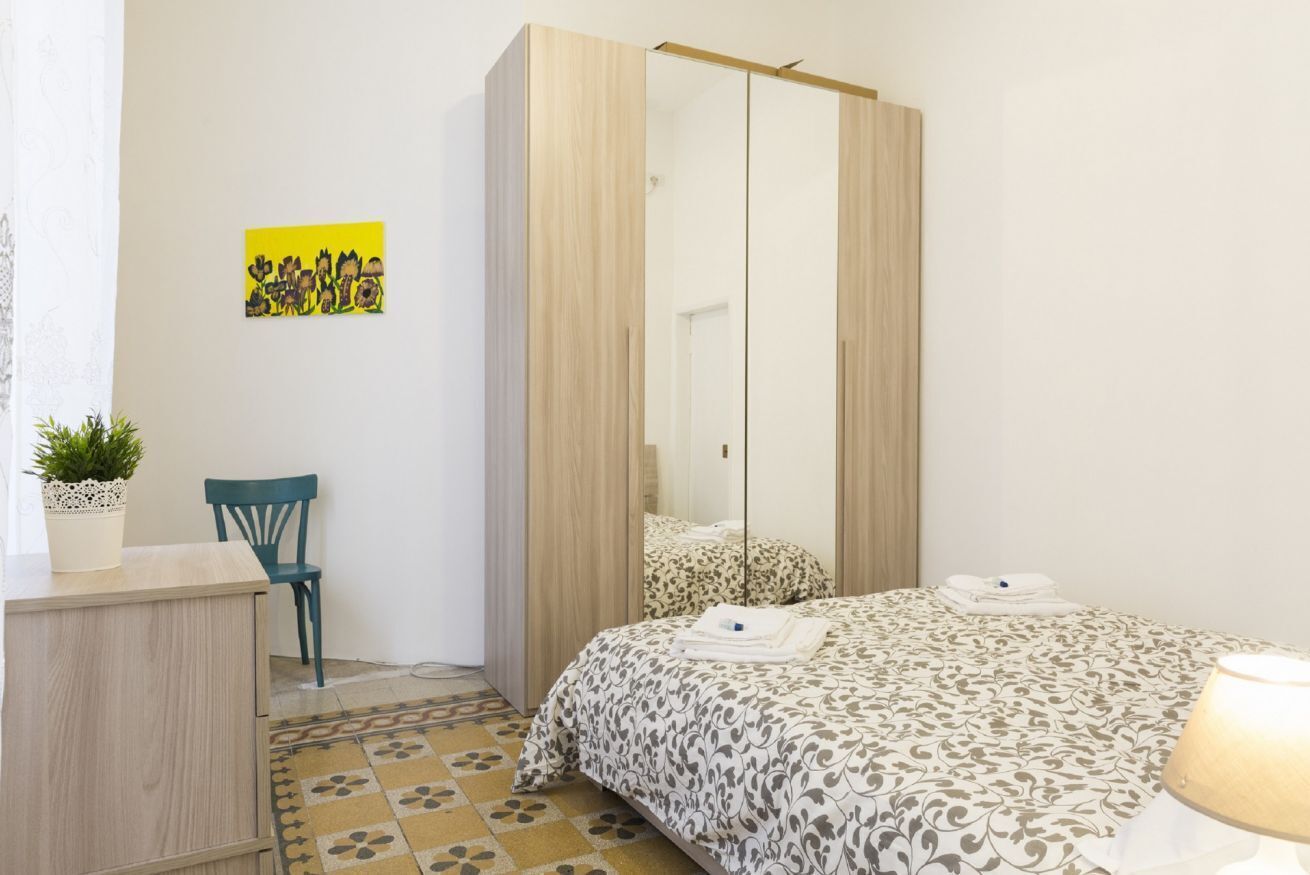 A bedroom with a bed, a wardrobe, a chair, a small table, and a potted plant.