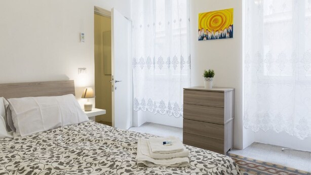 A bedroom with a bed, bedside table, lamp, dresser, and a window with curtains.