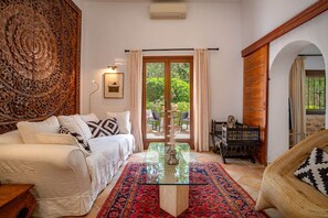 Una habitación con un sofá blanco, una mesa de centro de vidrio, un tapete estampado, un aparador de madera y vistas a través de un ventanal.