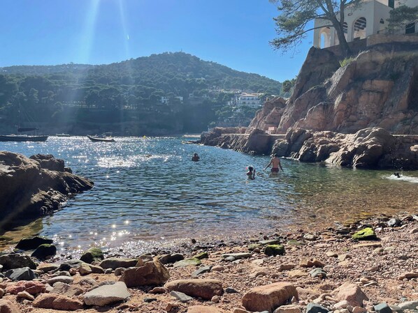 A rocky beach with clear water, boats, and a hill with buildings in the background.