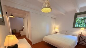 Een slaapkamer met een bed, twee nachtkastjes met lampen, een hanglamp, een witte deur en een raam met jaloezieën.