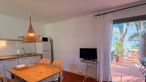 Een keuken met witte kastjes, een houten eettafel, rieten stoelen, een hanglamp, een televisie op een bijzettafeltje en een schuifpui die uitkomt op een terras buiten met uitzicht op palmbomen.