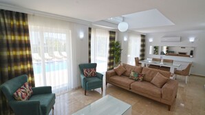 Een woonkamer met een bank, fauteuils, een glazen salontafel, en een eethoek met een tafel en stoelen.