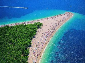 Ett flygbildsbild av en strand med en tät skog på ena sidan och en sandstrand med många parasoller och människor.
