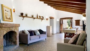 A living room with a stone fireplace, a sofa, a dining table with chairs, and a kitchen area visible through an open doorway. - CAL REI  Ref. VP34 (Pollensa / Pollença)