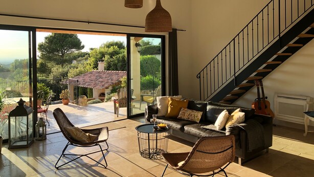 Ein großzügiges Wohnzimmer mit einem großen Glasfenster, das auf einen Balkon führt, einer Ledersofa, Korbstühlen und einer Treppe.