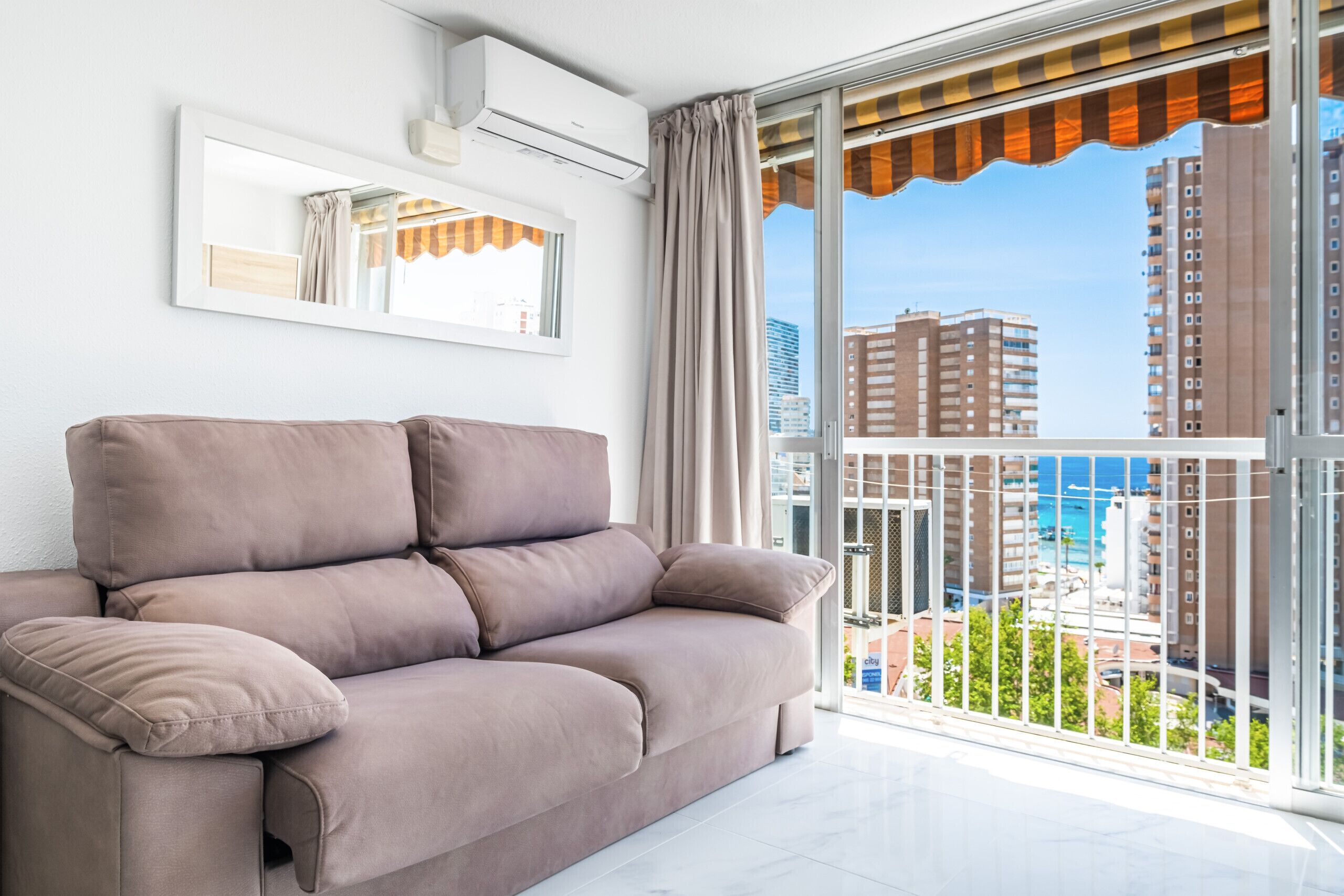 A modern living room with a beige sofa, a large window with a view of tall buildings and a blue sky.