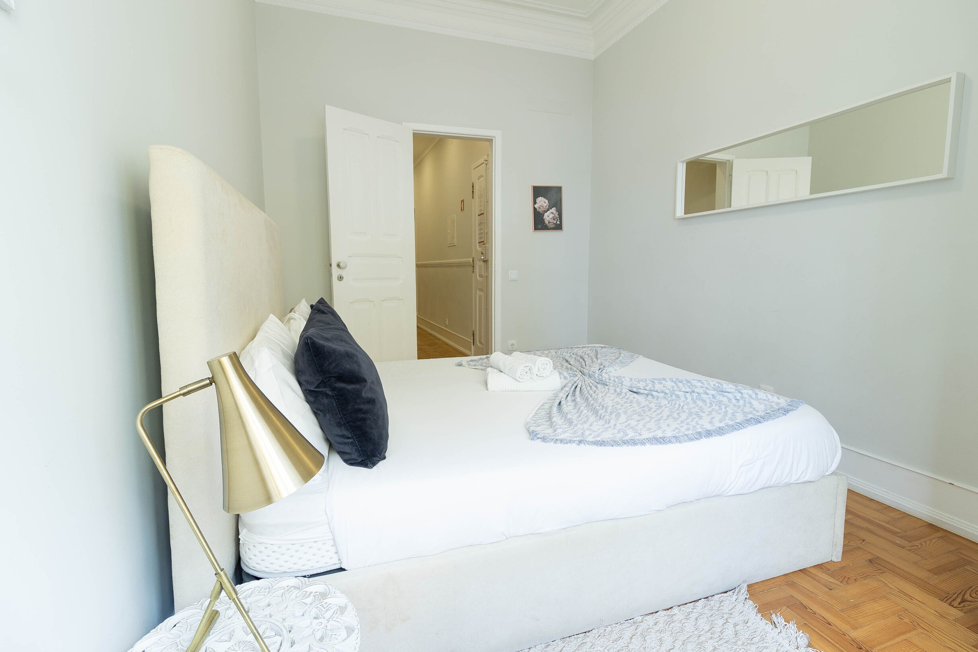 A bedroom with a bed, a bedside lamp, a mirror, and a door leading to another room.