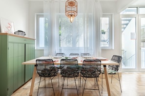 Uma área de refeições com uma mesa de madeira posta para seis pessoas, rodeada por cadeiras de estrutura de arame preto, um armário verde e uma janela grande com cortinas leves.